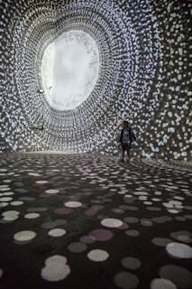 A person stands in a large, dark room surrounded by swirling patterns of white, circular lights projected on the walls and floor, creating an immersive tunnel-like visual experience.