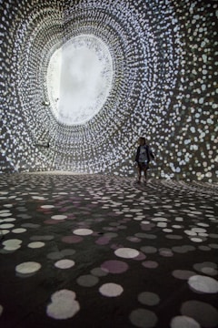 A person stands in a large, dark room surrounded by swirling patterns of white, circular lights projected on the walls and floor, creating an immersive tunnel-like visual experience.