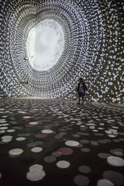 A person stands in a large, dark room surrounded by swirling patterns of white, circular lights projected on the walls and floor, creating an immersive tunnel-like visual experience.