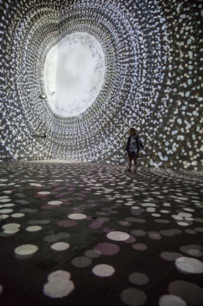 A person stands in a large, dark room surrounded by swirling patterns of white, circular lights projected on the walls and floor, creating an immersive tunnel-like visual experience.