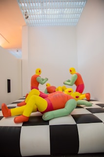 A group of colorful inflatable humanoid figures with yellow heads, green arms, and red bodies are seated and reclining on a large black and white checkered platform. They appear to be interacting in a playful and informal setting. The room is minimally decorated with white walls and a grid-patterned ceiling that allows ambient light to filter through.
