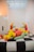 A group of colorful inflatable humanoid figures with yellow heads, green arms, and red bodies are seated and reclining on a large black and white checkered platform. They appear to be interacting in a playful and informal setting. The room is minimally decorated with white walls and a grid-patterned ceiling that allows ambient light to filter through.
