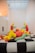 A group of colorful inflatable humanoid figures with yellow heads, green arms, and red bodies are seated and reclining on a large black and white checkered platform. They appear to be interacting in a playful and informal setting. The room is minimally decorated with white walls and a grid-patterned ceiling that allows ambient light to filter through.