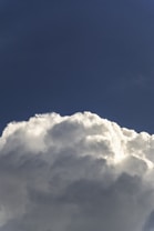 A large, fluffy cloud set against a deep blue sky. The cloud appears white with hints of gray shadows, suggesting a bright light source, possibly the sun.