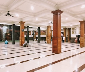 An interior space with large wooden pillars and polished tiled floors, where a few people are either standing or sitting on the ground in various postures. Ceiling fans are installed above, and there are intricate patterns on the walls. A podium or niche is visible at the far end.