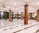 An interior space with large wooden pillars and polished tiled floors, where a few people are either standing or sitting on the ground in various postures. Ceiling fans are installed above, and there are intricate patterns on the walls. A podium or niche is visible at the far end.