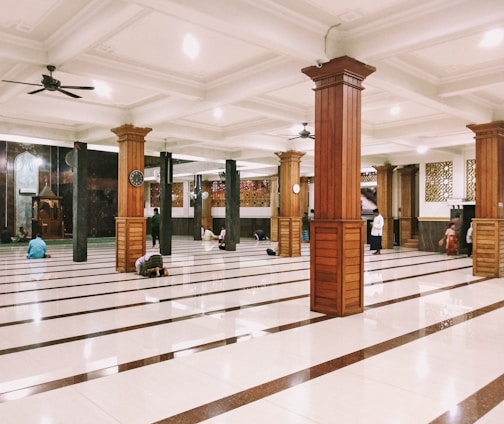 An interior space with large wooden pillars and polished tiled floors, where a few people are either standing or sitting on the ground in various postures. Ceiling fans are installed above, and there are intricate patterns on the walls. A podium or niche is visible at the far end.