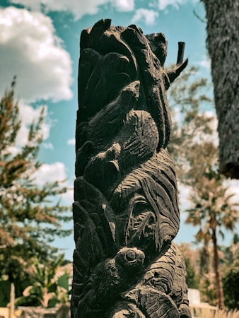 A tall, intricately carved wooden sculpture with swirling patterns stands outdoors. The background features a bright blue sky with fluffy white clouds and lush greenery, giving a natural and serene setting.