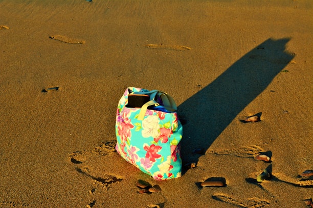 A close-up of a handcrafted beach bag resting on soft sand with sunlight filtering through palm leaves.