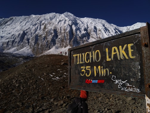 A rugged mountainous landscape with a large, snow-covered peak in the background. In the foreground, there is a weathered, metal sign indicating 'Tilicho Lake 35 Min.' with graffiti and stickers on it. A trail leads towards the mountain with a person carrying a backpack wearing an orange cap visible walking along the path.