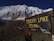 A rugged mountainous landscape with a large, snow-covered peak in the background. In the foreground, there is a weathered, metal sign indicating 'Tilicho Lake 35 Min.' with graffiti and stickers on it. A trail leads towards the mountain with a person carrying a backpack wearing an orange cap visible walking along the path.