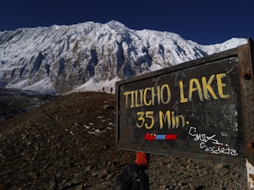 A rugged mountainous landscape with a large, snow-covered peak in the background. In the foreground, there is a weathered, metal sign indicating 'Tilicho Lake 35 Min.' with graffiti and stickers on it. A trail leads towards the mountain with a person carrying a backpack wearing an orange cap visible walking along the path.