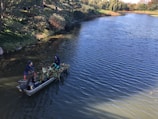 Two people are on a small boat in a scenic, tree-lined lake. They appear to be collecting or transporting aquatic plants, with the boat filled with greenery. The surrounding water is calm, reflecting the blue sky above, while trees with dense foliage line the banks.
