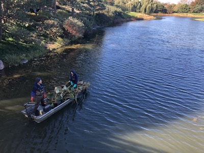 Two people are on a small boat in a scenic, tree-lined lake. They appear to be collecting or transporting aquatic plants, with the boat filled with greenery. The surrounding water is calm, reflecting the blue sky above, while trees with dense foliage line the banks.