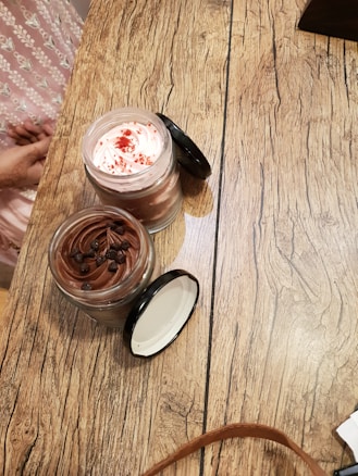 Two jars of dessert placed on a rustic wooden table. One jar contains a chocolate mousse topped with chocolate chips, while the other has a whipped cream topping with red sprinkles. The table's texture is a prominent feature, with visible wood grain patterns. A person wearing a pink patterned garment is partially visible in the background.