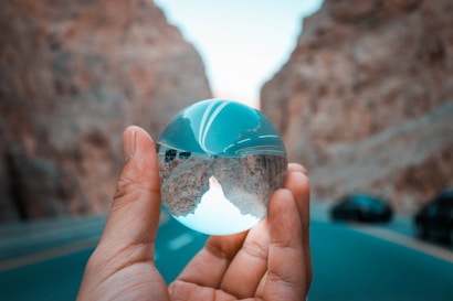 A hand holds a transparent crystal ball, reflecting an image of a winding road between steep rock cliffs. The background consists of blurred road and vehicles, creating a contrast with the clear reflection in the sphere.
