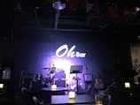 A dimly lit stage setup in a bar featuring musical instruments including a drum kit, microphone stands, and a guitar. There are tables with lighted candles in the foreground, adding to the ambient atmosphere. The wall in the background prominently displays a sign reading 'Oh Bar'.