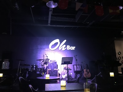 A dimly lit stage setup in a bar featuring musical instruments including a drum kit, microphone stands, and a guitar. There are tables with lighted candles in the foreground, adding to the ambient atmosphere. The wall in the background prominently displays a sign reading 'Oh Bar'.