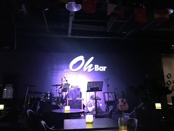 A dimly lit stage setup in a bar featuring musical instruments including a drum kit, microphone stands, and a guitar. There are tables with lighted candles in the foreground, adding to the ambient atmosphere. The wall in the background prominently displays a sign reading 'Oh Bar'.