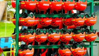 A collection of vibrant red helmets are organized neatly on metal shelves outdoors. Each helmet has black ventilation slits and features labels indicating some sort of branding or specification. The background includes a hint of greenery and other equipment.