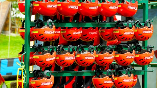 A collection of vibrant red helmets are organized neatly on metal shelves outdoors. Each helmet has black ventilation slits and features labels indicating some sort of branding or specification. The background includes a hint of greenery and other equipment.