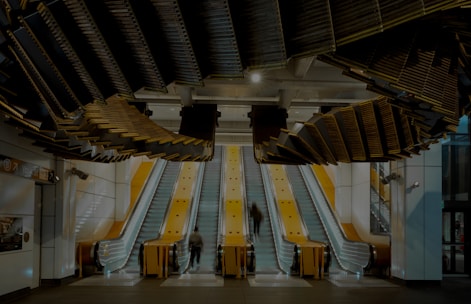 A modern architectural structure featuring three parallel yellow escalators leading upwards, surrounded by sleek, white walls. Above the escalators, an intricate and artistic ceiling installation adds a dramatic touch to the space. People are seen ascending on the escalators, captured in motion blur, adding dynamic movement to the otherwise static environment.