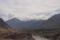 A rugged landscape featuring a range of towering mountains with snow-covered peaks under a partially cloudy sky. A winding river cuts through the barren land in the foreground, with patches of green vegetation visible along its banks.
