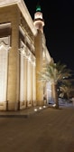A beautifully illuminated building with intricate architectural details stands against the night sky. The structure features tall, decorated columns and Arabic calligraphy near the top. A palm tree is situated nearby, and the presence of a minaret with a green light can be seen in the background.