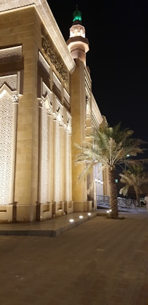 A beautifully illuminated building with intricate architectural details stands against the night sky. The structure features tall, decorated columns and Arabic calligraphy near the top. A palm tree is situated nearby, and the presence of a minaret with a green light can be seen in the background.