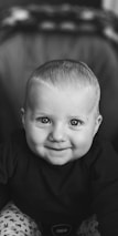 A smiling baby with bright eyes and short hair is dressed in a dark top and patterned pants. The black and white filter gives the image a classic and timeless feel.