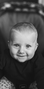 A smiling baby with bright eyes and short hair is dressed in a dark top and patterned pants. The black and white filter gives the image a classic and timeless feel.