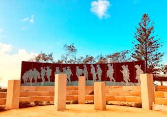 A memorial with metal silhouettes of soldiers marching, placed against a clear blue sky. The scene is surrounded by trees and large, light-colored stone pillars. The shadows of the soldiers are etched into a dark, curved panel supported by a brick wall.