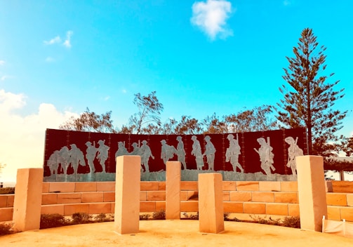 A memorial with metal silhouettes of soldiers marching, placed against a clear blue sky. The scene is surrounded by trees and large, light-colored stone pillars. The shadows of the soldiers are etched into a dark, curved panel supported by a brick wall.