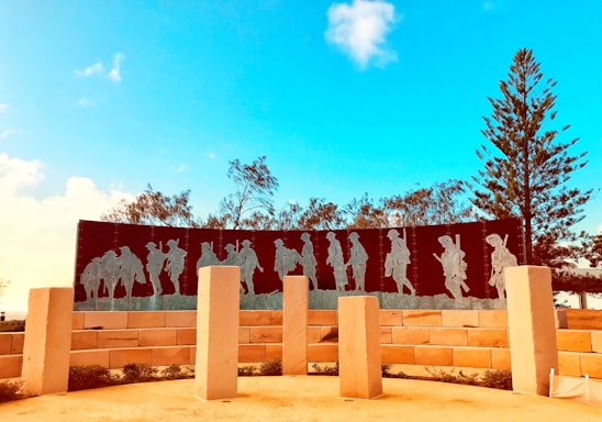 A memorial with metal silhouettes of soldiers marching, placed against a clear blue sky. The scene is surrounded by trees and large, light-colored stone pillars. The shadows of the soldiers are etched into a dark, curved panel supported by a brick wall.