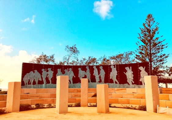A memorial with metal silhouettes of soldiers marching, placed against a clear blue sky. The scene is surrounded by trees and large, light-colored stone pillars. The shadows of the soldiers are etched into a dark, curved panel supported by a brick wall.