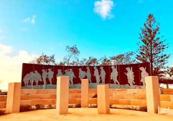 A memorial with metal silhouettes of soldiers marching, placed against a clear blue sky. The scene is surrounded by trees and large, light-colored stone pillars. The shadows of the soldiers are etched into a dark, curved panel supported by a brick wall.