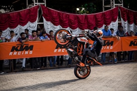 A motorcyclist performs a wheelie on an orange and black motorcycle in front of a crowd. The crowd is observing from behind a barrier with the KTM Racing logo. Red and white fabric drapes hang behind the spectators.