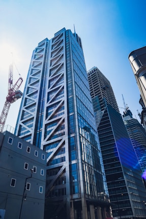 A modern skyscraper with a glass facade reflecting sunlight towers against a clear blue sky. The building features a distinctive architectural design with crisscrossing metal beams. Surrounding it are other high-rise buildings and a construction crane is visible, indicating ongoing development in an urban setting.