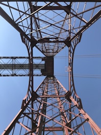 A perspective view of a large steel structure, likely a bridge or tower, showcasing intricate latticework against a clear blue sky. The structure's beams create geometric patterns that draw the eye upwards.