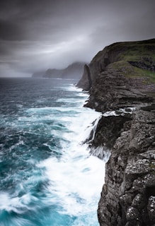 A dramatic coastal cliff under stormy skies with crashing salty waves below.