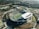 An aerial view of a large outdoor stadium with a green football field in the center. Surrounding the stadium are multiple sports fields, including a baseball field and what appears to be another practice field. The surrounding area has lush greenery and several buildings, likely part of a campus or sports complex. The scene is set in a suburban environment with residential neighborhoods and trees visible in the background.