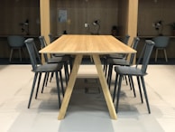 A tidy workspace featuring a long wooden table surrounded by several gray chairs. In the background, small workstations with chairs, desk lamps, and potted plants can be seen against a wooden wall. The setting is well-lit and organized, creating a modern and minimalist office environment.
