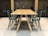 A tidy workspace featuring a long wooden table surrounded by several gray chairs. In the background, small workstations with chairs, desk lamps, and potted plants can be seen against a wooden wall. The setting is well-lit and organized, creating a modern and minimalist office environment.