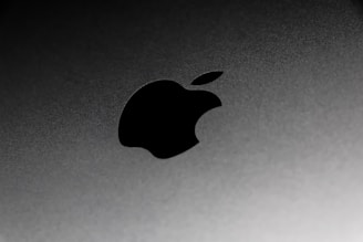 A close-up view of a metallic surface with a prominent black apple logo. The surface has a sleek and smooth texture with a subtle gradient of gray.