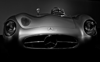 A vintage sports car with a sleek, aerodynamic design is captured from the front. The vehicle has a prominent Mercedes-Benz emblem on the grille, flanked by two rounded headlights. The smooth metal surface reflects light, highlighting its classic style.