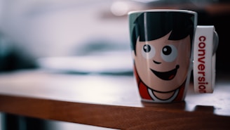A cartoon-character themed mug sits on a wooden table. The mug features a smiling face with large eyes and dark hair, and the handle has the text 'conversion' written vertically. The blurred background suggests an indoor setting with soft lighting.
