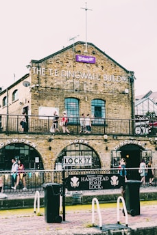 A historic brick building with the words 'The Dingwall Building' on the facade, featuring arched windows and doors. The structure is adjacent to a canal with lock gates labeled 'Lock 17'. People are walking and standing around the area, suggesting a lively atmosphere. There is a prominent sign reading 'Hampstead Road Lock 1'.
