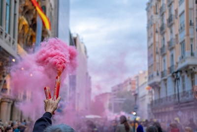 A hand holds two flares emitting pink smoke, set against an urban backdrop of ornate buildings. The street is crowded with people, and there is a Spanish flag hanging from one of the buildings.