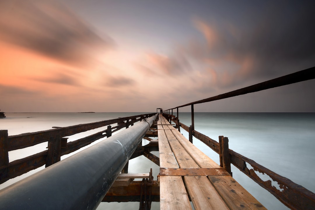 brown wood plank bridge with gray metal pipe above water under gray clouds