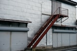 A metal staircase painted in rust-red rises along a large building with a silvery-grey exterior. Below, several closed roller shutters line the building, each in a light grey color. Above the shutters, there are small, rectangular windows with frosted or textured glass. The surface of the building appears to be made from large metal panels, some showing signs of wear or slight dents.